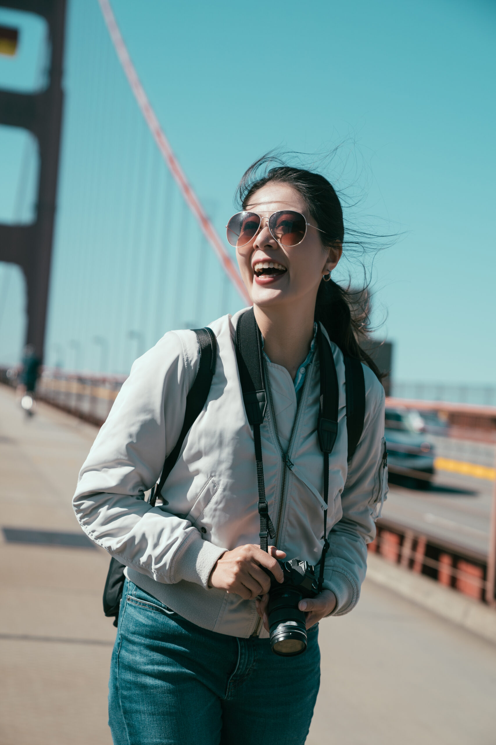 cheerful tourist photographer laughing holding professional camera standing under sunshine on red golden gate bridge in san francisco on pedestrian with cars dirving pass on road in background.