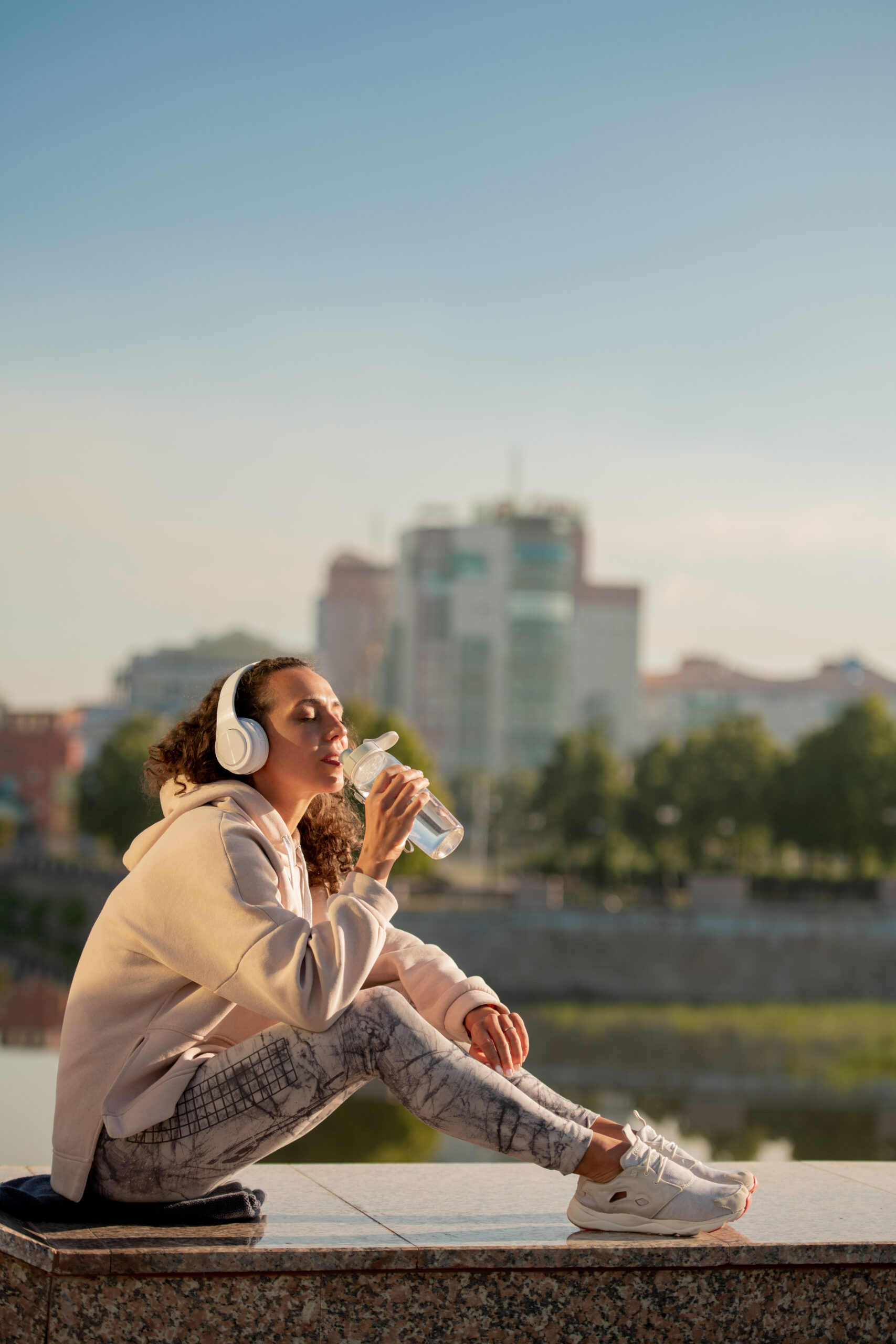 Restful thirsty female in sportswear drinking water from bottle while sitting against riverside after outdoor training and enjoying music